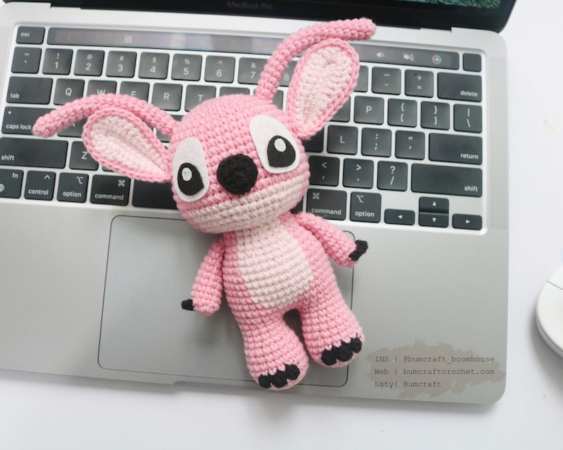 May include: Pink crocheted amigurumi toy with large black eyes and a black nose. The toy is sitting on a laptop keyboard. The text "INS | @bumcraft_boonhouse Web | bumcraftcrochet.com Estyl Bumcraft" is visible on the laptop.