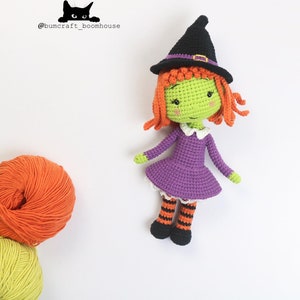 Puede incluir: Una mu&ntilde;eca de bruja de crochet con piel verde, cabello naranja, un vestido morado y medias a rayas negras y naranjas. La mu&ntilde;eca lleva un sombrero de bruja negro con una banda morada.