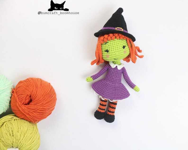 Puede incluir: Una mu&ntilde;eca de bruja de crochet con piel verde, cabello naranja, un vestido morado y medias a rayas negras y naranjas. La mu&ntilde;eca lleva un sombrero de bruja negro con una banda morada.