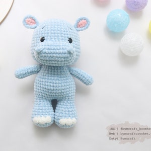 May include: A light blue crocheted hippopotamus toy with pink ears and white feet. The toy is sitting on a white surface with colorful string lights in the background.