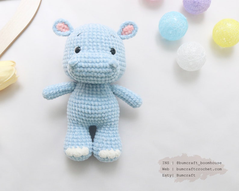 May include: A light blue crocheted hippopotamus toy with pink ears and white feet. The toy is sitting on a white surface with colorful string lights in the background.
