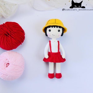 May include: A crocheted doll wearing a yellow hat, white shirt, red skirt, and red shoes. The doll is sitting on a white surface with two balls of yarn, one red and one pink.