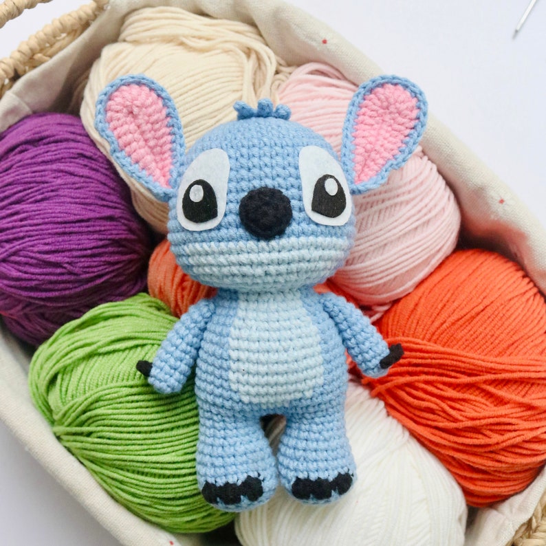 May include: A blue crocheted Stitch character toy with pink ears and black eyes. The toy is surrounded by balls of yarn in various colors, including purple, green, pink, orange, and white.