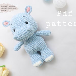 May include: A light blue crocheted hippopotamus toy with white accents on the ears and feet. The toy is lying on a white surface with yellow tulips in the background. The text "Pdf pattern" is written in white on the surface.