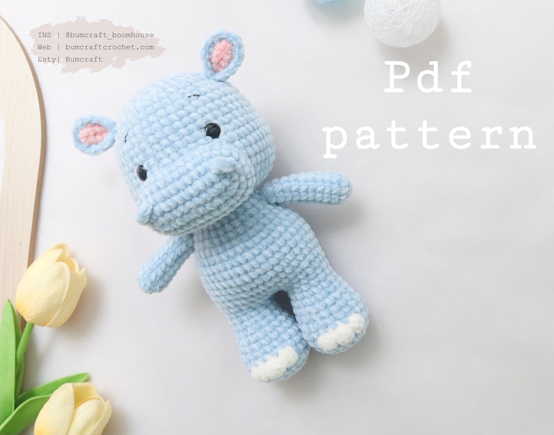 May include: A light blue crocheted hippopotamus toy with white accents on the ears and feet. The toy is lying on a white surface with yellow tulips in the background. The text "Pdf pattern" is written in white on the surface.