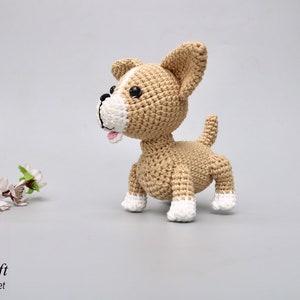 May include: A crocheted tan and white dog toy with black eyes. The dog is standing on a white surface.