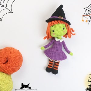 Puede incluir: Una mu&ntilde;eca de bruja de crochet con piel verde, cabello naranja, un vestido morado y medias rayadas de color negro y naranja. La mu&ntilde;eca lleva un sombrero de bruja negro con una banda marr&oacute;n. La mu&ntilde;eca est&aacute; sentada sobre un fondo blanco.