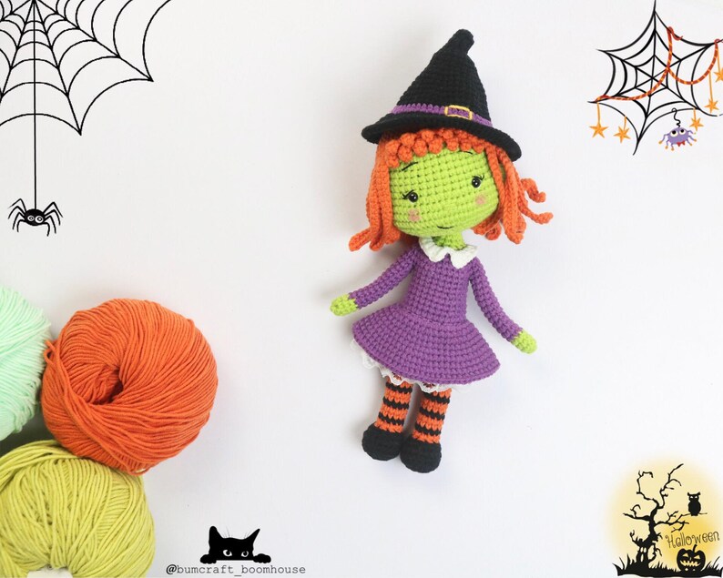 Puede incluir: Una mu&ntilde;eca de bruja de crochet con piel verde, cabello naranja, un vestido morado y medias rayadas de color negro y naranja. La mu&ntilde;eca lleva un sombrero de bruja negro con una banda marr&oacute;n. La mu&ntilde;eca est&aacute; sentada sobre un fondo blanco.
