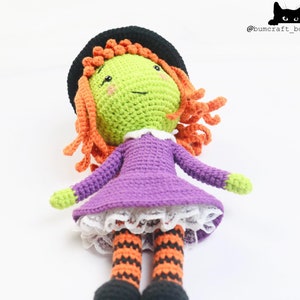 Puede incluir: Una mu&ntilde;eca de bruja de crochet con pelo naranja, un vestido morado y medias a rayas negras y naranjas. La mu&ntilde;eca lleva un sombrero negro y tiene una cara verde con un cuello blanco.
