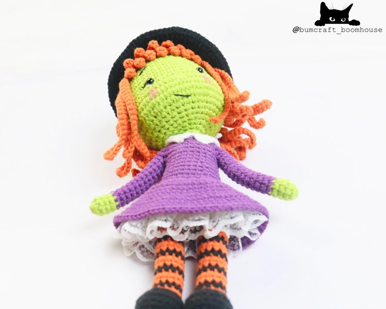 Puede incluir: Una mu&ntilde;eca de bruja de crochet con pelo naranja, un vestido morado y medias a rayas negras y naranjas. La mu&ntilde;eca lleva un sombrero negro y tiene una cara verde con un cuello blanco.