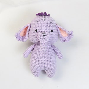 Könnte beinhalten: Ein gehäkeltes lila Elefanten-Plüschtier mit rosa Ohren und einer lila Blume auf dem Kopf.