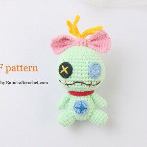 May include: A green crocheted plush toy with pink ears and a yellow antenna. The toy has a red collar, blue eyes, and a stitched mouth. The toy is sitting on a white background. PDF pattern by Bumcraftcrochet.com