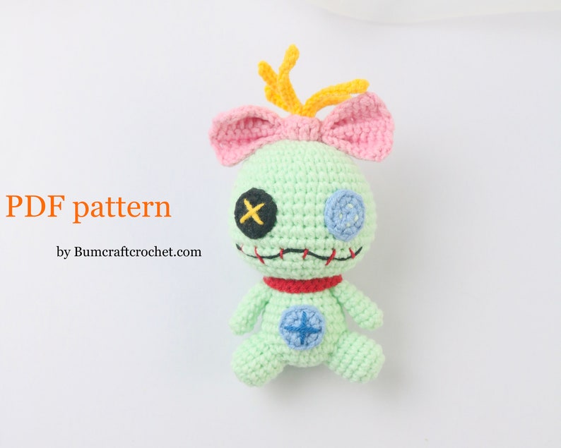 May include: A green crocheted plush toy with pink ears and a yellow antenna. The toy has a red collar, blue eyes, and a stitched mouth. The toy is sitting on a white background. PDF pattern by Bumcraftcrochet.com