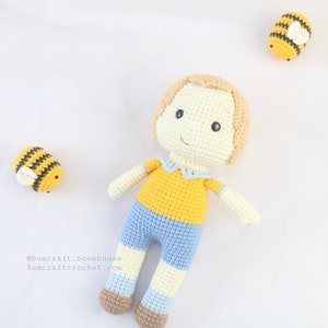 May include: A crocheted doll with blonde hair, wearing a yellow shirt, blue shorts, and white socks. The doll is lying on a white surface with two crocheted bees nearby.