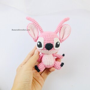 May include: Pink crocheted amigurumi toy with black eyes and a black nose. The toy is in the shape of a cartoon character with large ears and a small body. The toy is being held in a person's hand.