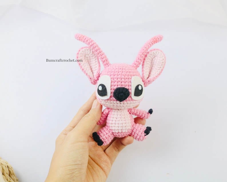 May include: Pink crocheted amigurumi toy with black eyes and a black nose. The toy is in the shape of a cartoon character with large ears and a small body. The toy is being held in a person's hand.