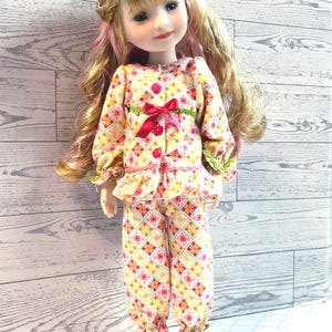 May include: A doll with long blonde hair and pink highlights, wearing a floral patterned pajama set. The pajamas have a yellow, pink, and orange diamond pattern, a red bow, and green trim. The doll has a fair complexion and is posed in a standing position.