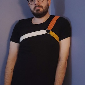 May include: Black t-shirt with a white diagonal stripe and a crochet harness in orange and yellow. The harness is connected with a silver ring. The person is wearing blue jeans and glasses.