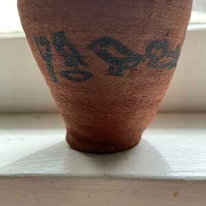 Puede incluir: Una pequeña taza de cerámica de color terracota con una parte superior ensanchada y una base estrecha. La taza está decorada con un diseño de pájaros negros alrededor de la parte superior. La taza está sobre una superficie blanca.
