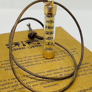 May include: A clear glass vial pendant with a gold-colored cap, containing a rolled papyrus scroll with hieroglyphs. The pendant hangs on a brown leather cord. The vial is resting on a brown paper with text.
