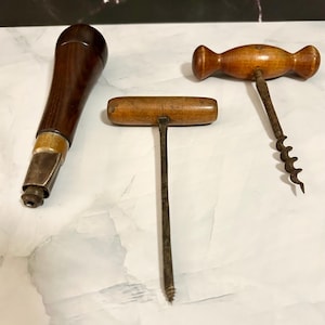 May include: Three antique corkscrews. One has a dark wooden handle with a metal base. Another is a simple T-shaped corkscrew with a wooden handle. The third has a wooden handle and a metal corkscrew. All are vintage wine tools.