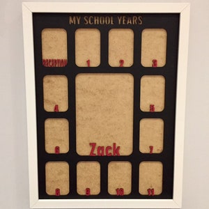 May include: A white rectangular frame containing a black mat with the text MY SCHOOL YEARS at the top. The mat features twelve small rectangular openings labeled with red numbers from 1 to 11 and the word RECEPTION, plus a large central opening with the name Zack in red.