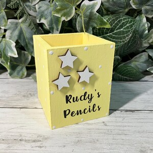 May include: A square yellow wooden pencil holder featuring white polka dots, three white star-shaped embellishments, and the black text "Rudy's Pencils" printed on the front side. The container has an open top and a flat base.