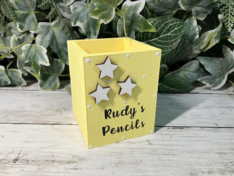 May include: A square yellow wooden pencil holder featuring white polka dots, three white star-shaped embellishments, and the black text "Rudy's Pencils" printed on the front side. The container has an open top and a flat base.