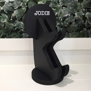 K&ouml;nnte beinhalten: Schwarzer Holzst&auml;nder mit einer runden Oberseite, auf der "JODIE" in wei&szlig;en Buchstaben steht. Der St&auml;nder ist in Form einer Person mit erhobenen Armen gestaltet.