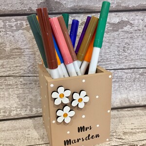 May include: A square tan pen holder with white polka dots, three white flower embellishments with yellow centers, and the black text "Mrs Marsden" printed on the front. The holder is filled with a variety of colorful felt-tip markers.