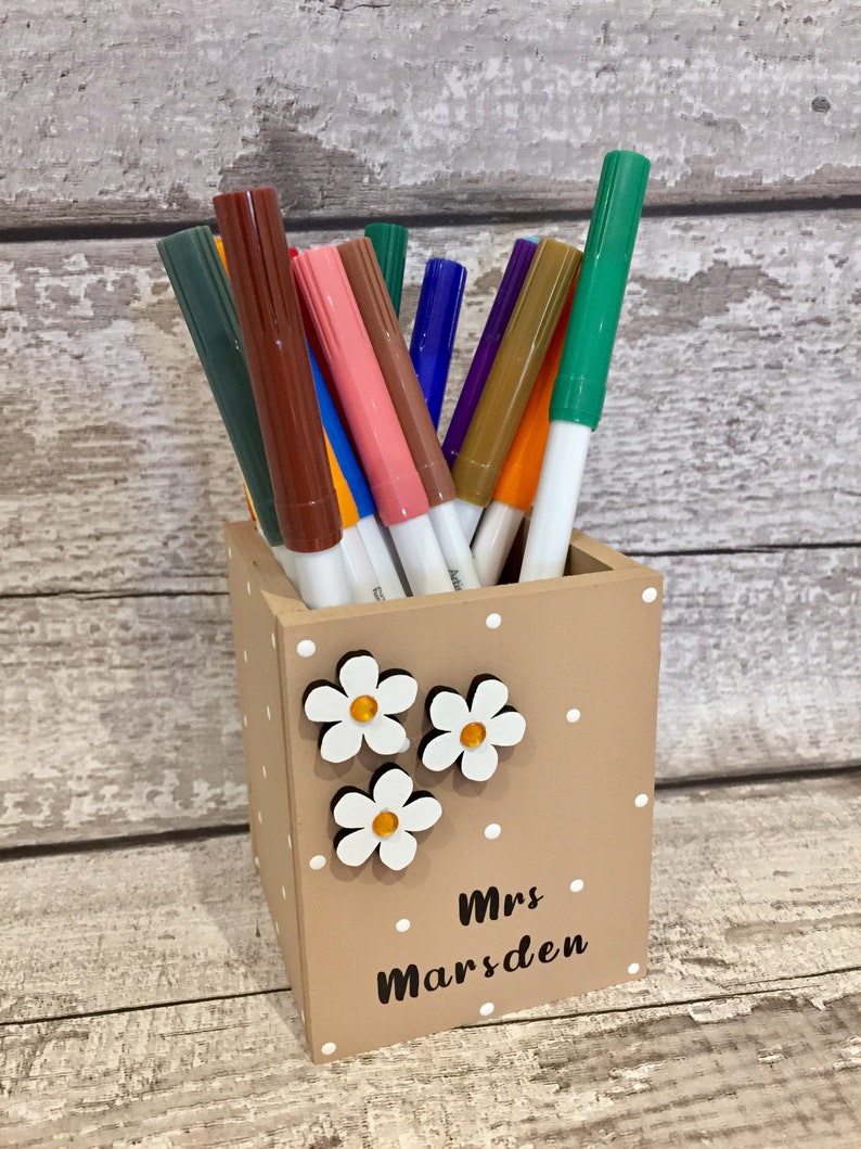 May include: A square tan pen holder with white polka dots, three white flower embellishments with yellow centers, and the black text "Mrs Marsden" printed on the front. The holder is filled with a variety of colorful felt-tip markers.
