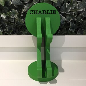 K&ouml;nnte beinhalten: Gr&uuml;ner, h&ouml;lzerner Kopfh&ouml;rerst&auml;nder mit runder Basis und einer runden Oberseite, auf der "CHARLIE" in schwarzen Buchstaben steht.