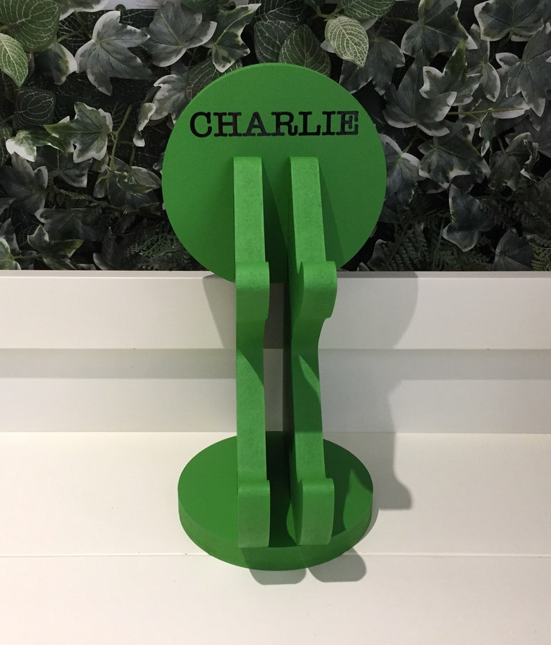 K&ouml;nnte beinhalten: Gr&uuml;ner, h&ouml;lzerner Kopfh&ouml;rerst&auml;nder mit runder Basis und einer runden Oberseite, auf der "CHARLIE" in schwarzen Buchstaben steht.