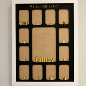 May include: A white rectangular picture frame with a black mat featuring the text MY SCHOOL YEARS at the top. It includes twelve small photo slots labeled Reception through 11 in gold, plus a larger central slot with the name Lainey in gold.