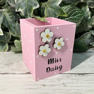 May include: A square pink wooden pencil holder featuring a white polka dot pattern. The front side is decorated with three white flower cutouts with yellow centers and the black text "Miss Daisy" printed in a script font on the lower section.