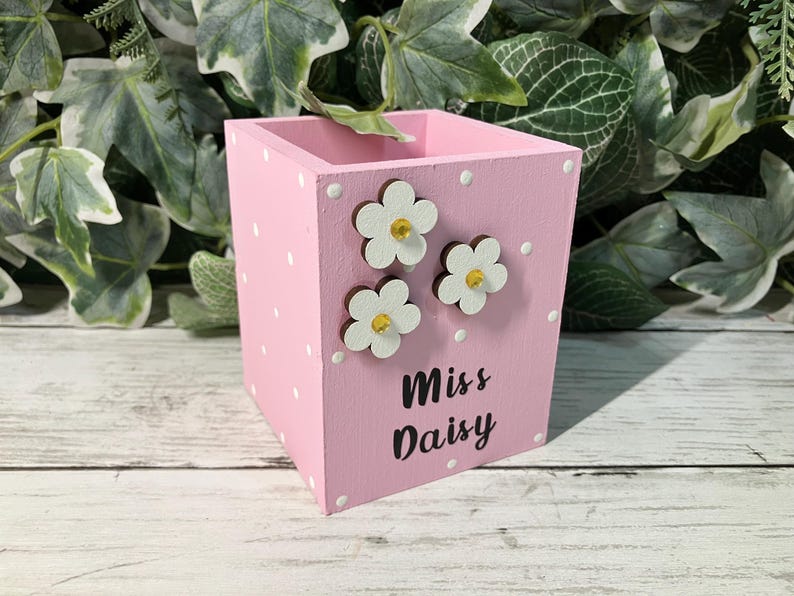 May include: A square pink wooden pencil holder featuring a white polka dot pattern. The front side is decorated with three white flower cutouts with yellow centers and the black text "Miss Daisy" printed in a script font on the lower section.
