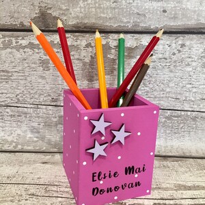 May include: A square pink pencil holder featuring white polka dots, three light purple star cutouts, and the black printed text Elsie Mai Donovan. The container holds six colored pencils in orange, red, yellow, green, dark red, and brown.