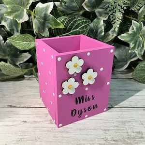 May include: A square pink wooden pencil holder featuring white polka dots, three white flower embellishments with yellow centers, and the black text "Miss Dyson" printed on the front side of the container for desk organization.