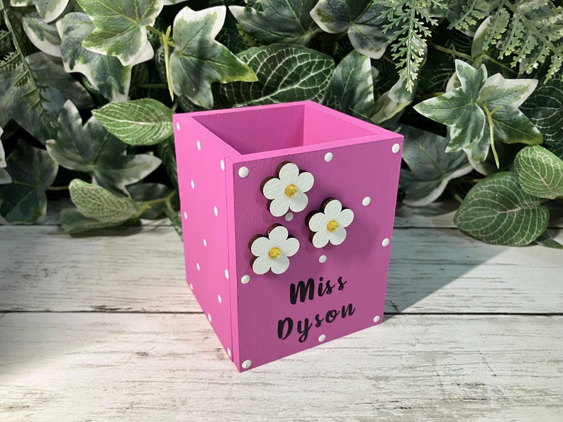 May include: A square pink wooden pencil holder featuring white polka dots, three white flower embellishments with yellow centers, and the black text "Miss Dyson" printed on the front side of the container for desk organization.