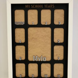 May include: A white-framed rectangular display board titled MY SCHOOL YEARS in black text. It features twelve tan photo slots labeled with silver numbers from Reception to 11, plus a larger central slot with the silver name Myla displayed below.