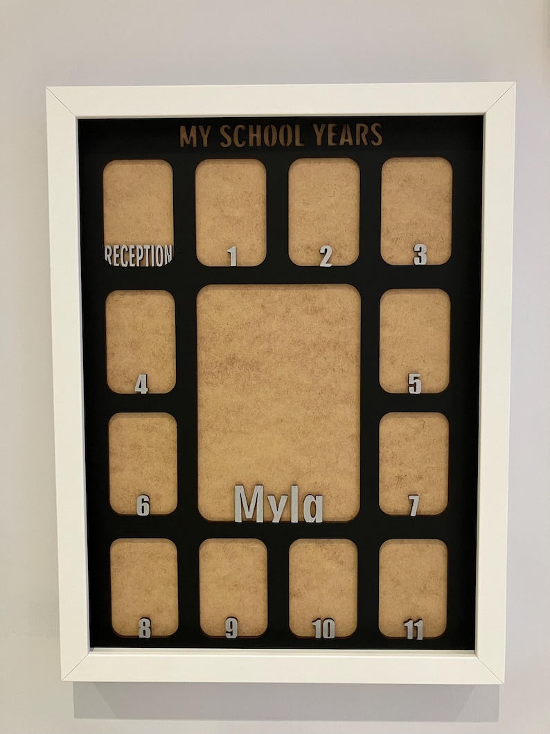 May include: A white-framed rectangular display board titled MY SCHOOL YEARS in black text. It features twelve tan photo slots labeled with silver numbers from Reception to 11, plus a larger central slot with the silver name Myla displayed below.