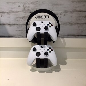 May include: A white gaming headset stand with the name "Jesse" printed on the front. Two white Xbox controllers are attached to the stand.
