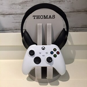 K&ouml;nnte beinhalten: Wei&szlig;er Xbox-Controller und schwarzer Kopfh&ouml;rer auf einem St&auml;nder mit dem Namen "THOMAS" darauf.