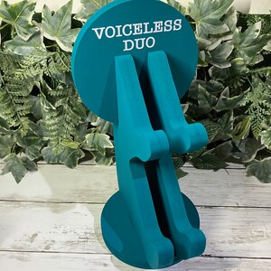 K&ouml;nnte beinhalten: Tealblauer Holz-Telefonst&auml;nder mit zwei Steckpl&auml;tzen f&uuml;r Telefone. Der St&auml;nder hat eine runde Basis mit dem Aufdruck "VOICELESS DUO".