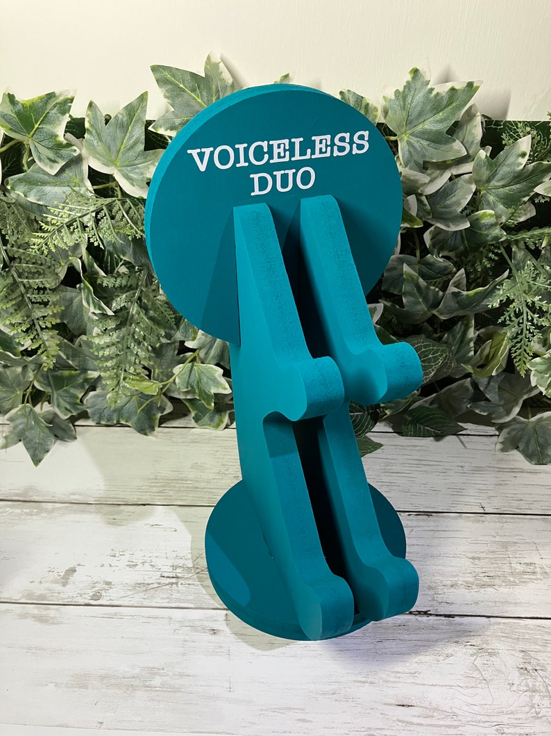 K&ouml;nnte beinhalten: Tealblauer Holz-Telefonst&auml;nder mit zwei Steckpl&auml;tzen f&uuml;r Telefone. Der St&auml;nder hat eine runde Basis mit dem Aufdruck "VOICELESS DUO".