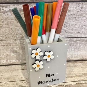 May include: A square gray wooden pencil holder with white polka dots, decorated with three white flower cutouts featuring orange centers. The text "Mrs Marsden" is printed in black on the front. The holder is filled with various colored markers.