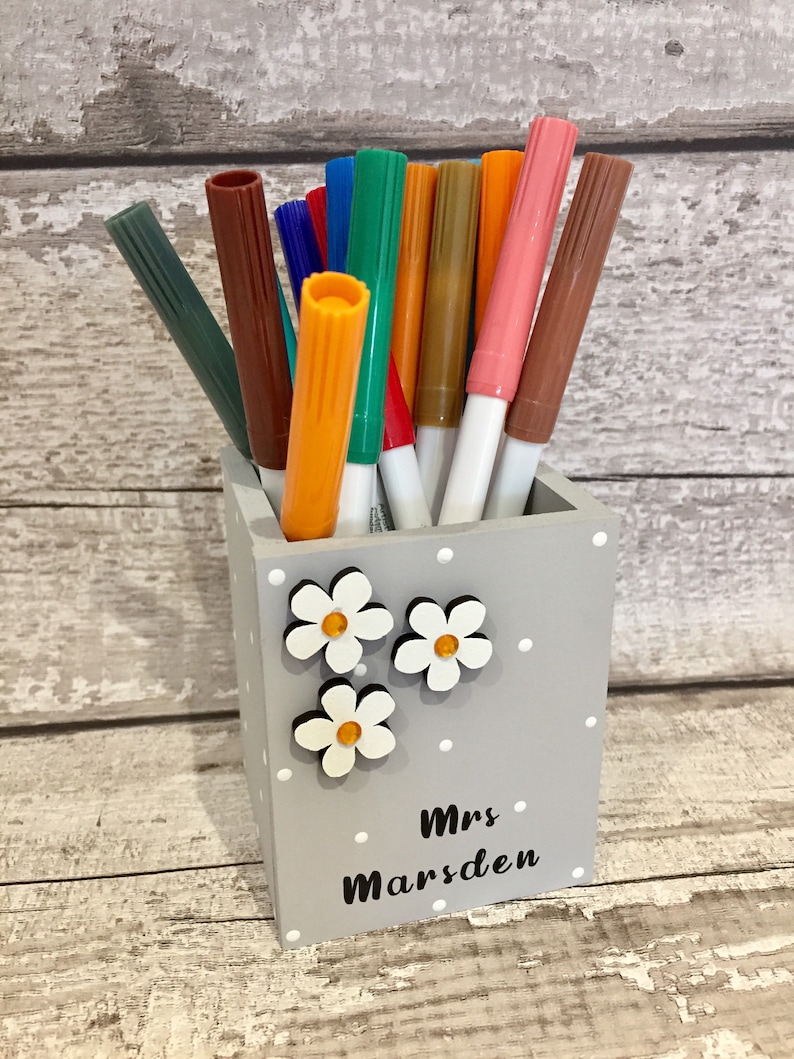 May include: A square gray wooden pencil holder with white polka dots, decorated with three white flower cutouts featuring orange centers. The text "Mrs Marsden" is printed in black on the front. The holder is filled with various colored markers.