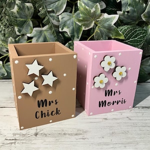 May include: Two square wooden pen holders, one tan with white star cutouts and the text Mrs Chick, and one pink with white flower cutouts and the text Mrs Morris. Both containers feature white polka dots painted on the sides and front surfaces.