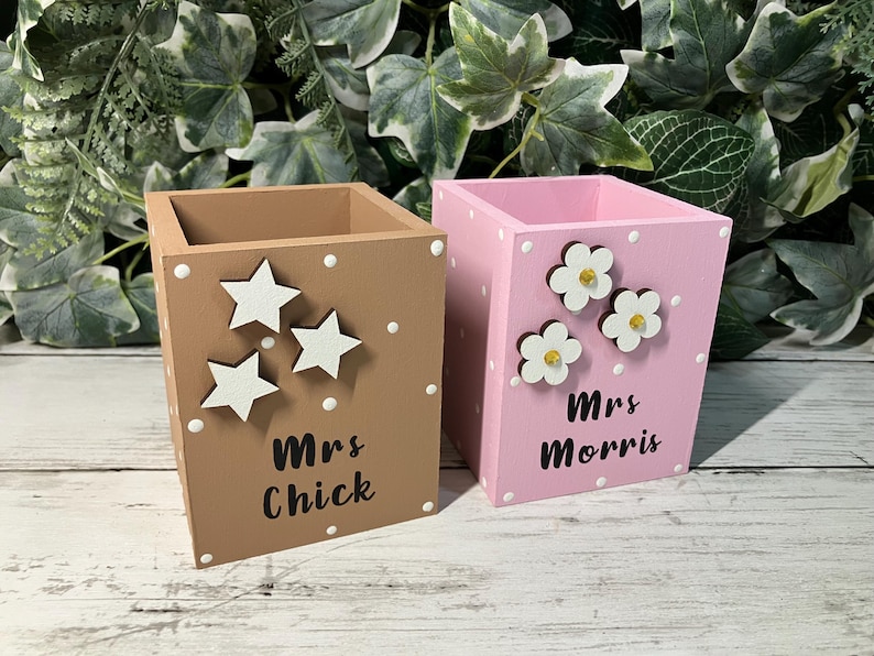 May include: Two square wooden pen holders, one tan with white star cutouts and the text Mrs Chick, and one pink with white flower cutouts and the text Mrs Morris. Both containers feature white polka dots painted on the sides and front surfaces.