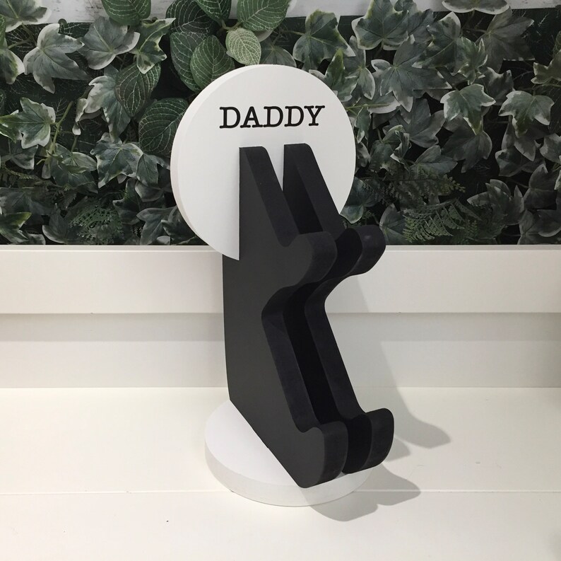 K&ouml;nnte beinhalten: Ein wei&szlig;er Holzst&auml;nder mit einer schwarzen Silhouette einer Person, die ein Schild mit der Aufschrift "DADDY" h&auml;lt.